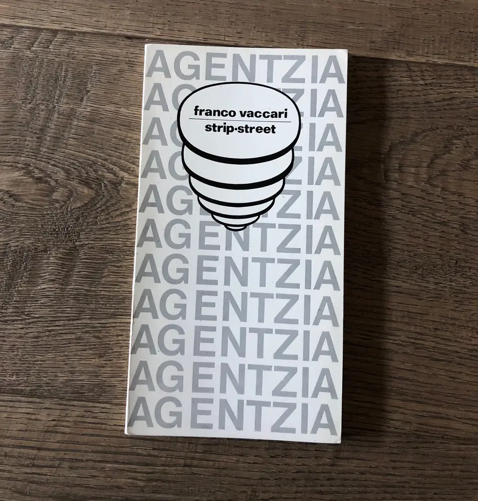 Franco Vaccari’s ‘strip-street’ book lies on a wooden surface, showing its minimalist cover with repeated ‘AGENTZIA’ lettering.